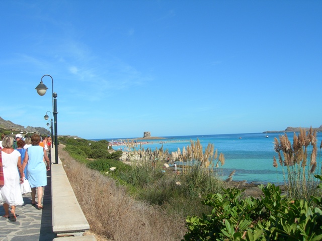 Il gruppo percorre il lungomare verso la spiaggia che li accogliera'. Sotto il mare azzurrissimo chiama, mentre in lontananza fa capolino la torre di osservazione.